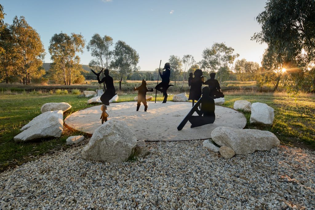 Photo of the artwork "Family Gathering" by Michael Quinn from the Yindyamarra Sculpture Walk in Albury. Features seven Indigenous figures cut out of metal, in silhouette, gathered in a circle as if engaged in ceremony. 